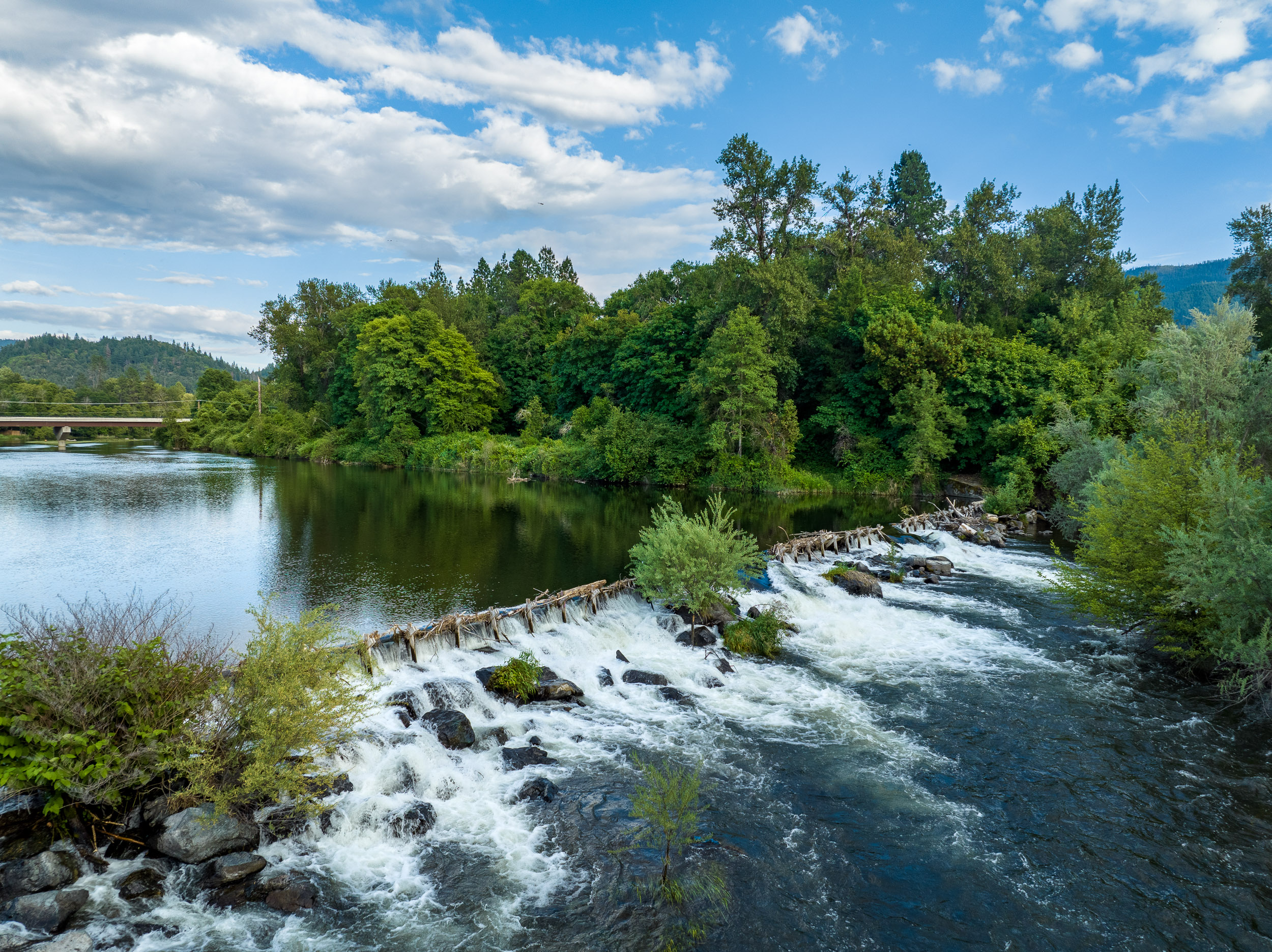 A weir and diversion structure spanning the Rogue River in Josephine County, Oregon, with forested banks and a bridge in the distance.